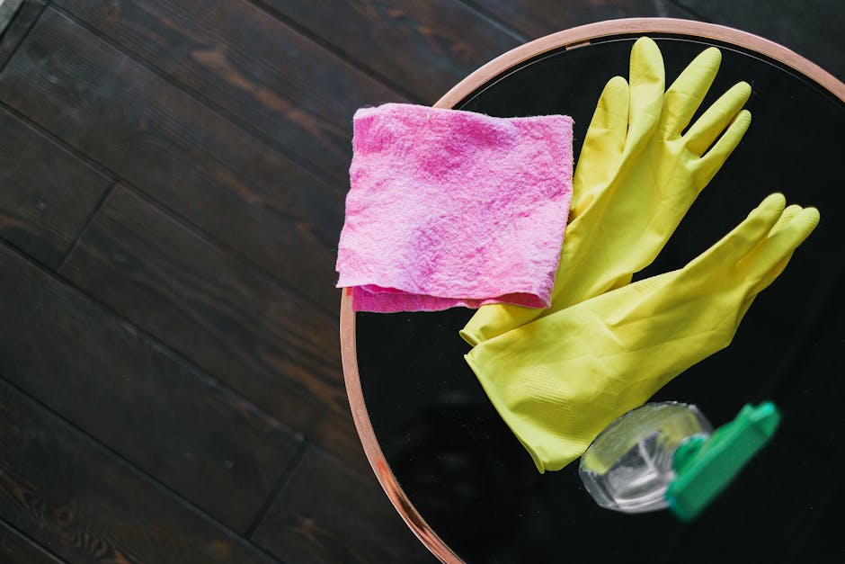 A close-up view of a round black table with a light wooden rim, positioned on a dark wooden floor. On the table, there is a neatly folded pink microfiber cloth, a pair of bright yellow rubber gloves,